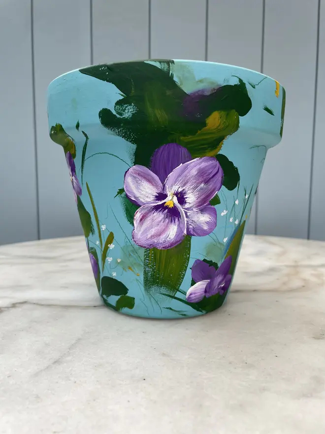 Unique hand-painted terracotta pot featuring purple pansy flowers on a pale blue background
