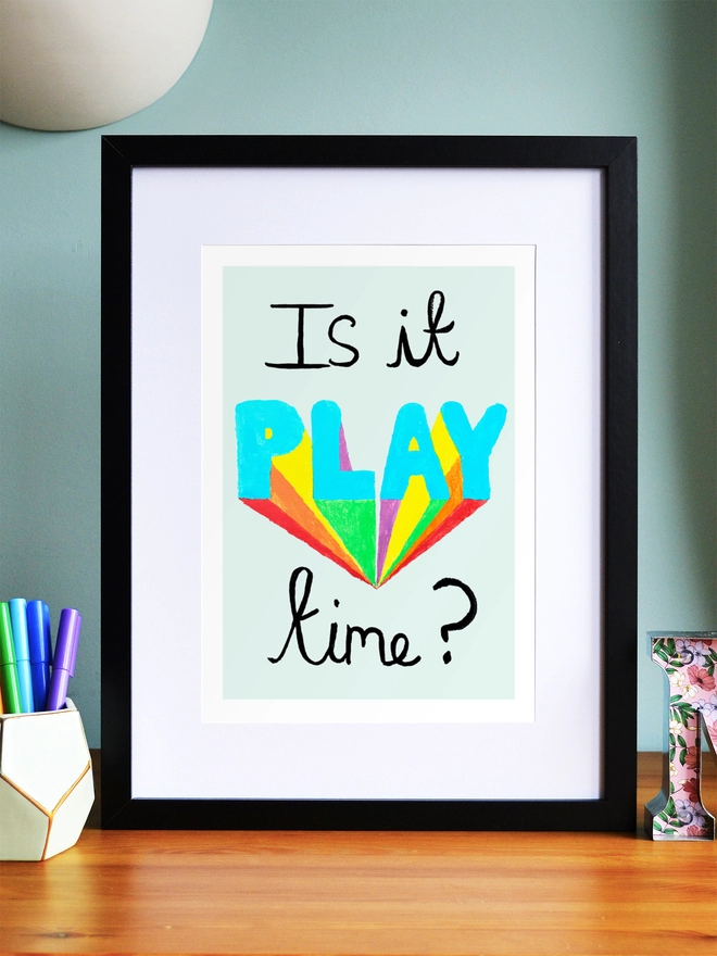 Art print saying 'Is it play time?' in a black frame on a child's desk