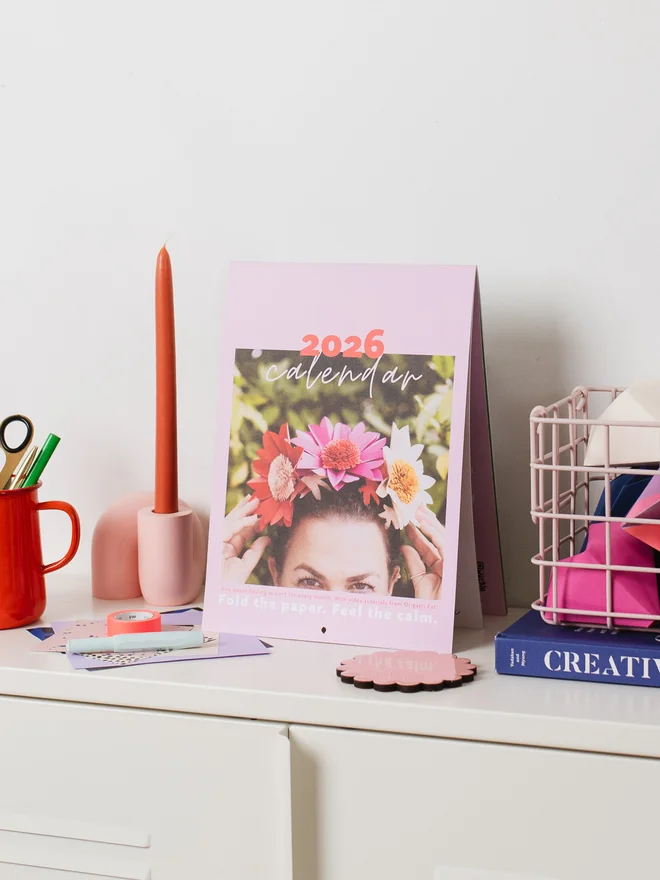 Calendar balanced on a cabinet, with a jug, candle, book & basket styled around it.