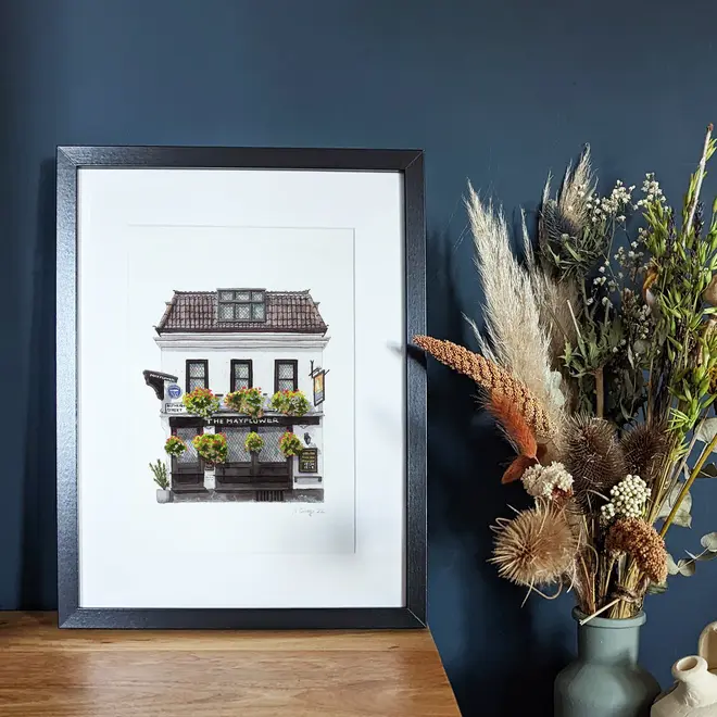 Rotherhithe The Mayflower Pub Giclée Print (Unframed), a print of a pub sitting in a black frame against a wooden surface against a navy blue backdrop. 