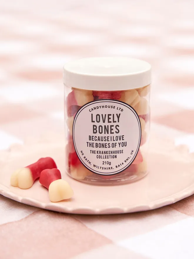 A clear jar with a white lid filled with jelly white and red bones labelled Lovely Bones on a pink frill plate places on a pink checkered tablecloth