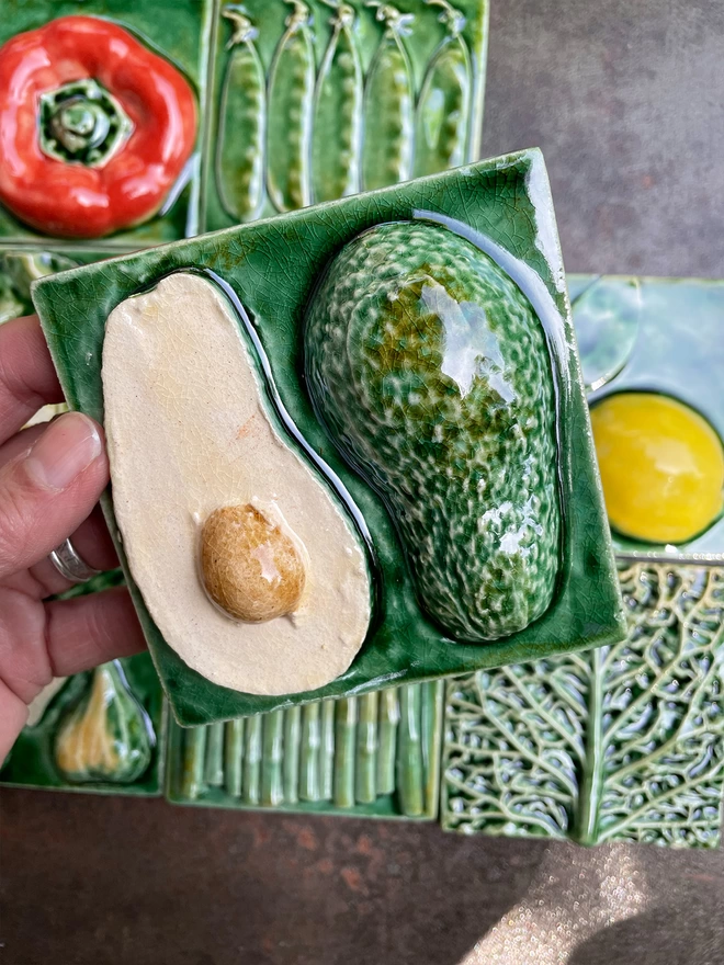 Avocado tile - square, 3D, realistic and glossy. The avocado - cut in half to reveal the brown pip inside – is on a lush green background. Other fruit and vegetable tiles in the series are on display in the background: lemon, fig, savoy cabbage, pear, mange tout, red capsicum pepper, garlic, asparagus.