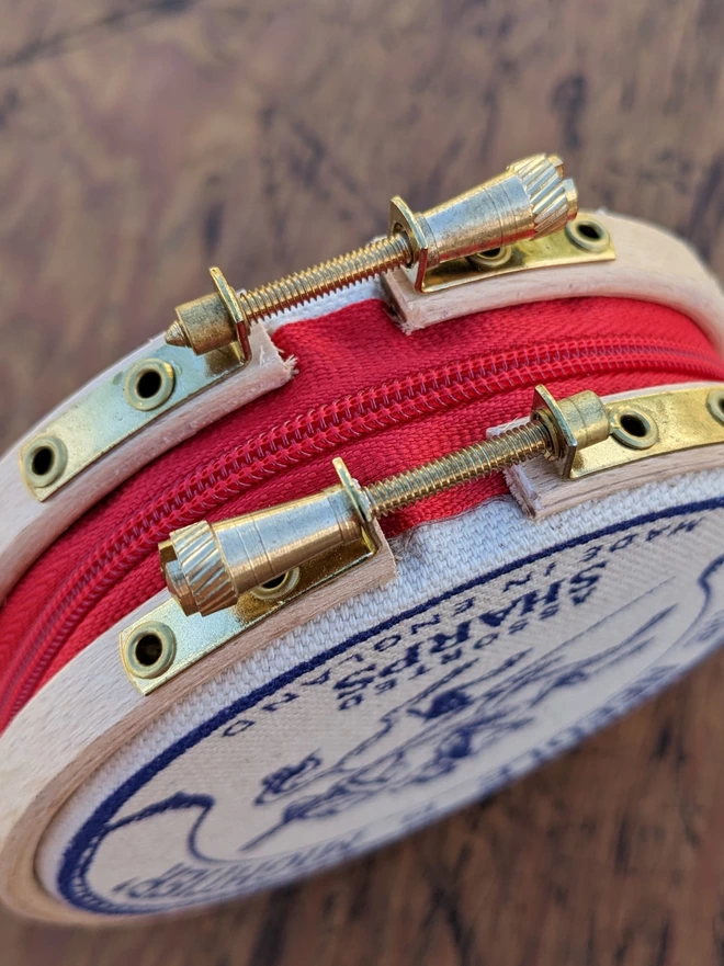 close up of red zip and gold clasps on the royal blue coloured 'the needle is mightier' embroidery hoop needle book