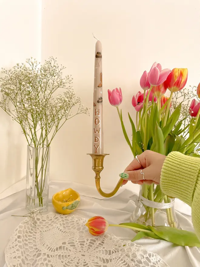 'Howdy' Personalised Hand Painted Candle, a woman placing the candle on a table top accompanied by flowers. It's design can be seen wrapping around the stick. 