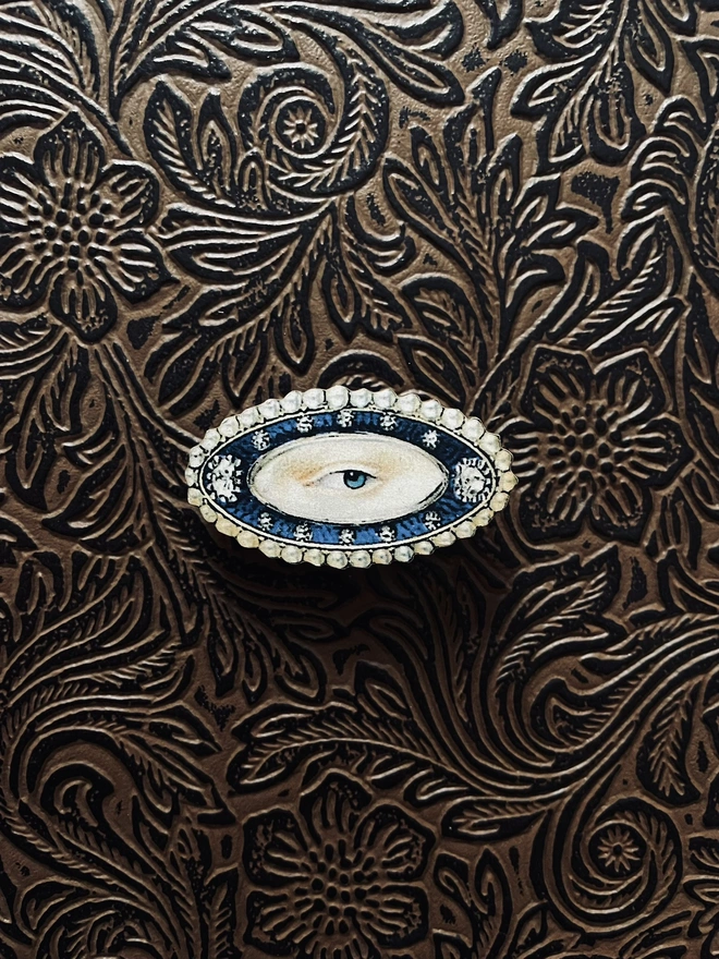 a lovers eye brooch, illustration of a lovers eye surrounded by jewels is printed to wood, laser cut, and has brass pin on reverse