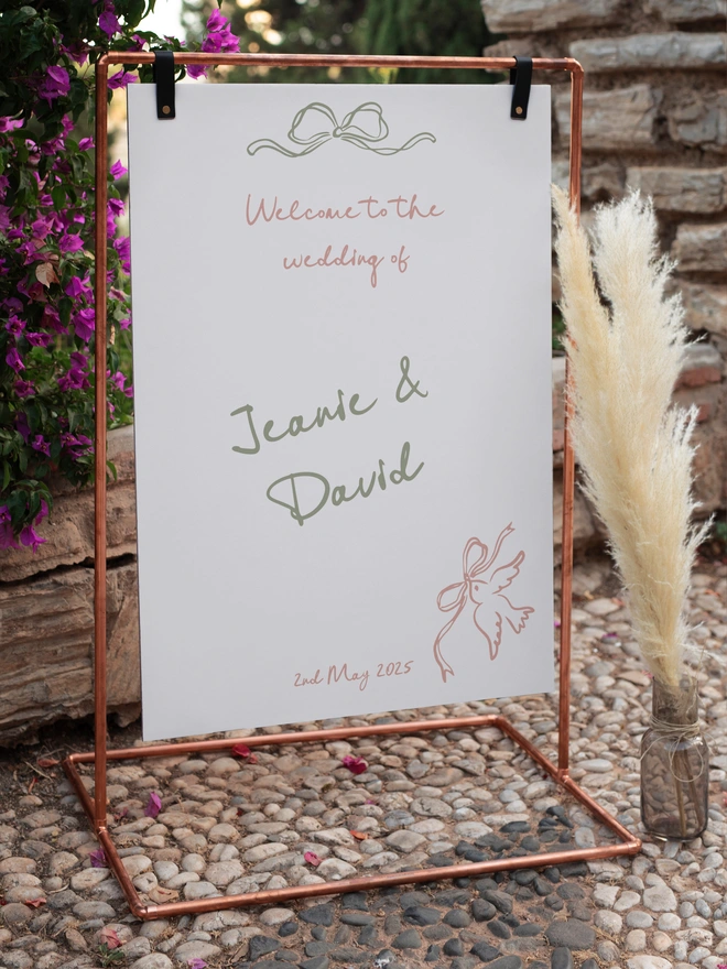 a coper pipe frame with a white board hanging in the middle from two black leather straps, the board is white and has the wording in dusky pink lettering, welcome to the wedding of and in Khaki lettering there is the names Jeanie and David plus in Khaki colour there is a whimsical bow at the top and in the right hand corner a bird carrying a bow in dusky pink