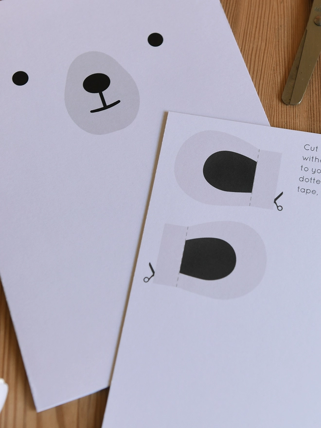 Two white polar bear greetings cards lay on a wooden table, one shows to front with the face, the other shows the back with the ears.