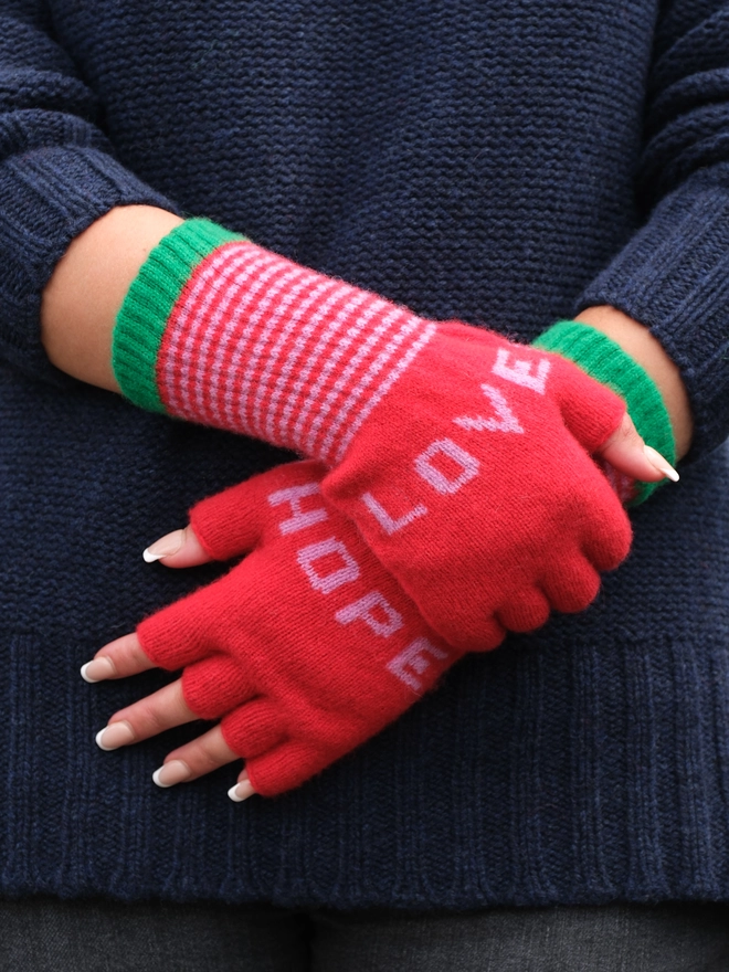 red fingerless gloves with pink words . one hand says LOVE and the other hand says HOPE . there is also a pink striped cuff and a green edge at the top of the cuff. They are warm for wrists and hands but keep the fingers free for phone and computer use.a fun and practical gift