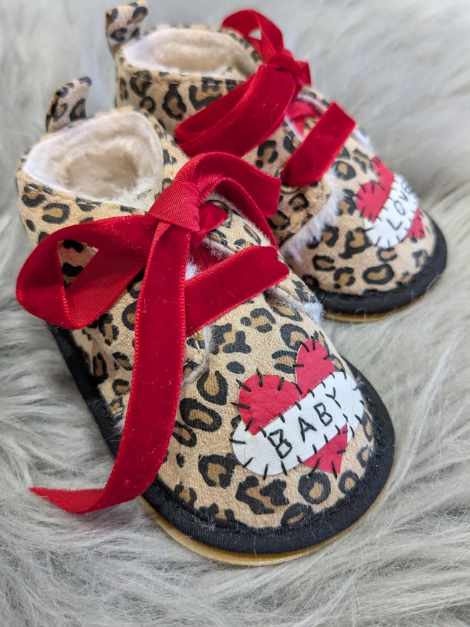 A pair of leopard print baby booties, lined with fur and with red velvet ribbon laces. A red heart with a white scroll adorns each booty. One says BABY, the other, LOVE in black stitches