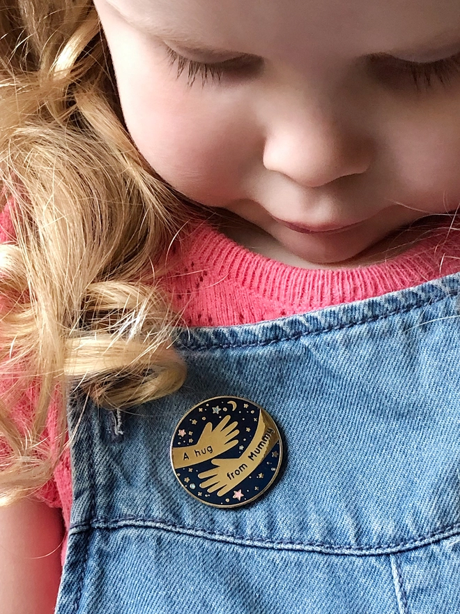 A small child has a navy blue and gold enamel pin badge with a hugging arms design and the words "A hug from Mummy" pinned to her blue dungarees.