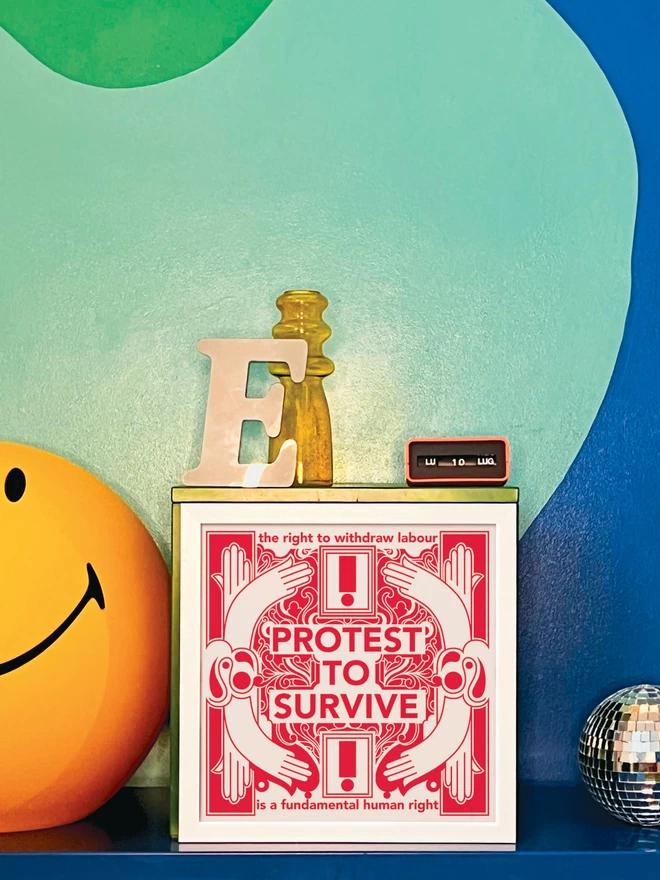The bold red and white print with "Protest to survive" written at the centre, as well as “the right to withdraw labour” at the top, and “is a fundamental human right” at the bottom, is in a white frame propped against a turquoise and dark blue wall. Next to the frame is a disco ball,a letter ‘E’ ornament, a yellow glass vase, an orange Italian plastic calendar showing the date as ‘LU 10 LUG’ and a large light up yellow Smiley lamp.
