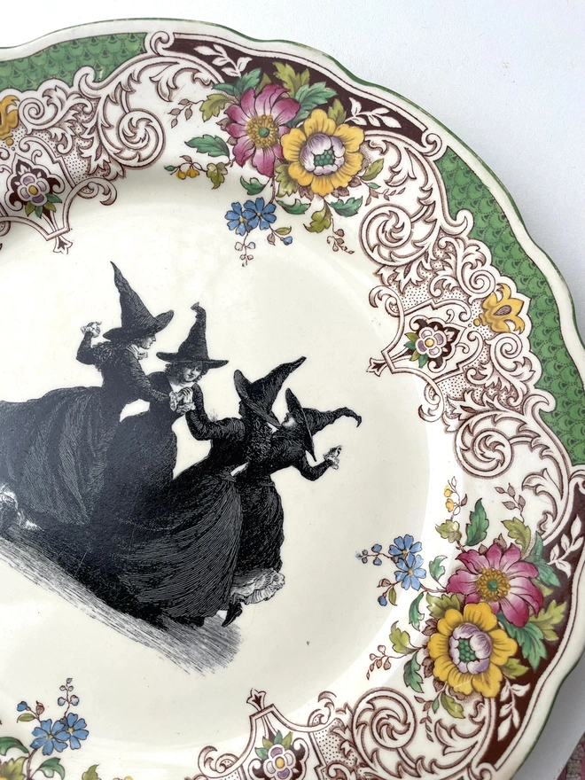 a vintage plate with an ornate floral border in the centre is a witch's coven, the four of them are hand in hand dancing in a circle