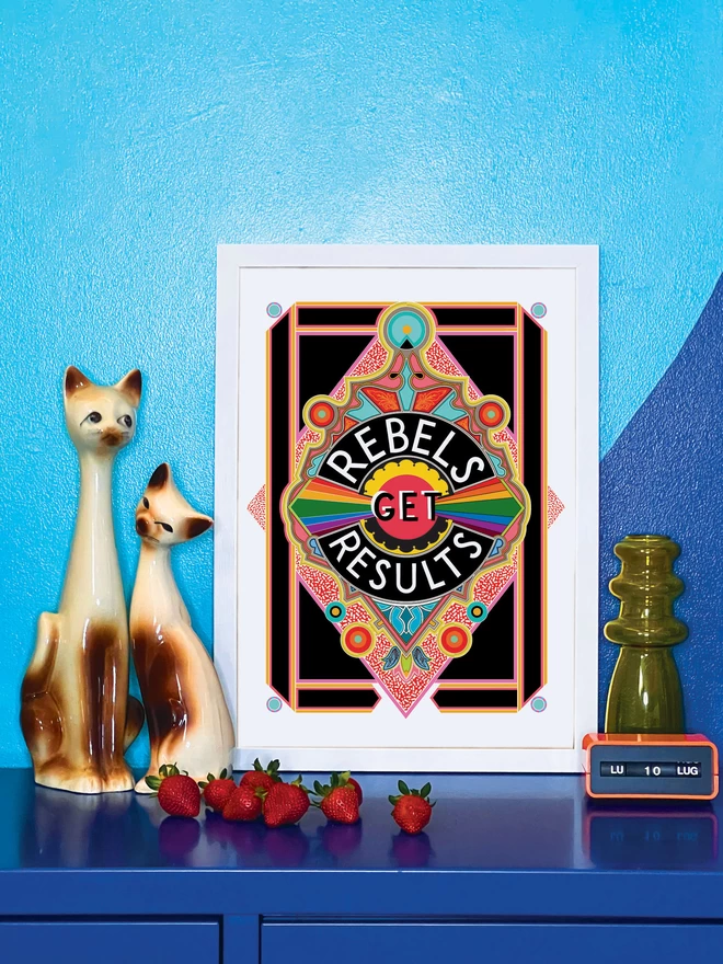 Rebels Get Results is written in white on a black background at the centre of this vibrant, abstract portrait illustration, with a black background and rainbows emitting from the centre, and multi-coloured detailing. The picture is in a white frame, against a turquoise and blue wall resting on a blue cabinet. Next to the picture are two cat ornaments, some ripe strawberries, a yellow glass vase and an orange Italian plastic calendar showing the date as ‘LU 10 LUG’.