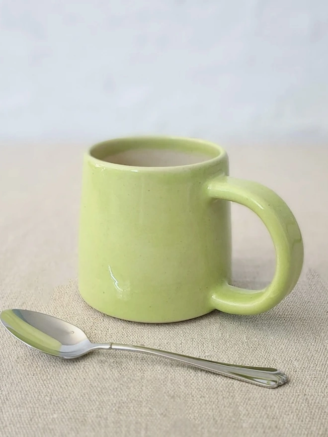 Ceramic Standard Mug, a colourful ceramic mug sitting on a plain surface against a grey backdrop. It is accompanied by a spoon. 