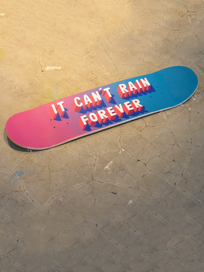A skateboard that has a spray painted pink and blue background and 3d typography that reads It Can’t Rain Forever in pink and white lies on a concrete floor. Artwork by Survival Techniques