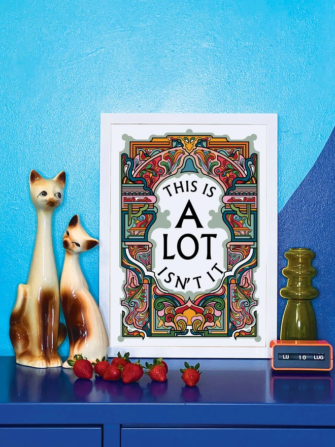 This is A Lot, Isn’t it is written in black on a white background at the centre of this bold, symmetrical portrait illustration. The picture is in a white frame, against a turquoise and blue wall resting on a blue cabinet. Next to the picture are two cat ornaments, some ripe strawberries, a yellow glass vase and an orange Italian plastic calendar showing the date as ‘LU 10 LUG’.