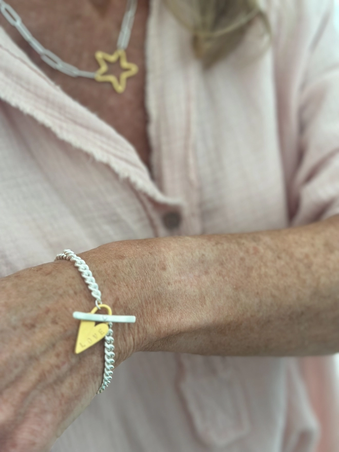 model wears a sterling silver chunky curb bracelet with silver t bar fastener and personalised gold chunky heart padlock charm