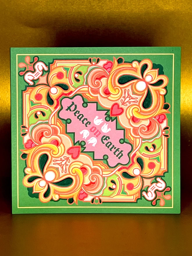 An abstract Christmas card design with Peace on Earth at the centre with drawings of two doves above and two below, surrounded by a multi-coloured design on a green background. The card features greens, pinks and oranges and is displayed in front of a gold backdrop. 