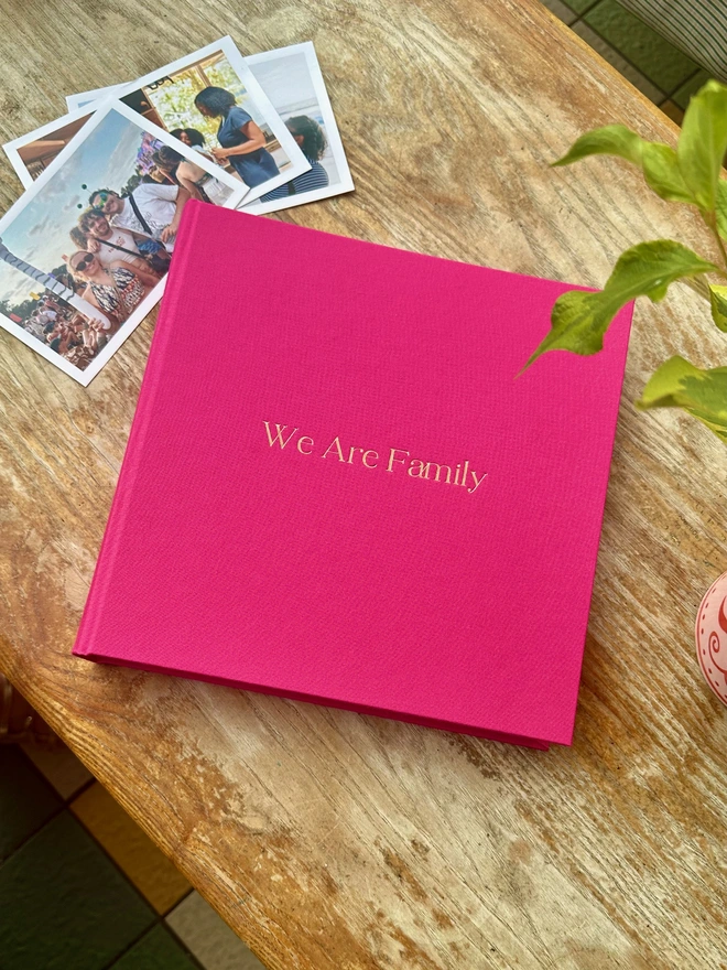 A hot pink photo album printed with 'We Are Family' in gold lettering sits on a wooden surface, next to a pile of photos.
