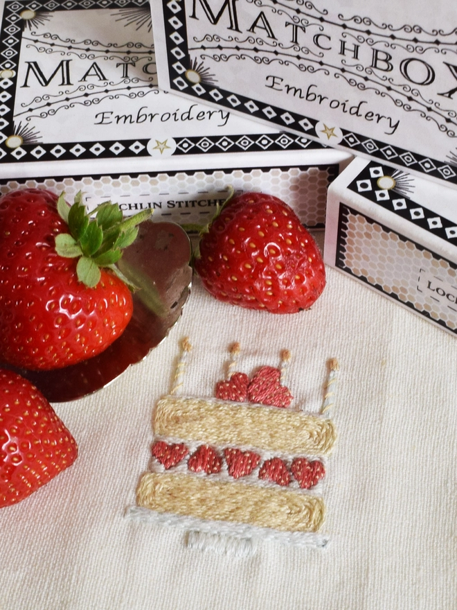 Mini embroidery of a classic Victoria sponge cake with strawberries and cream, topped with 4 candles, displayed with fresh home grown strawberries and the matchbox packaging in the background.