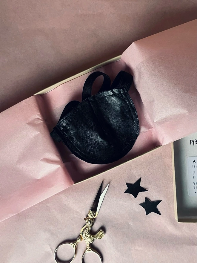 Black leather eye patch placed on pink tissue paper in brown box, gold scissors and black stars placed next to it
