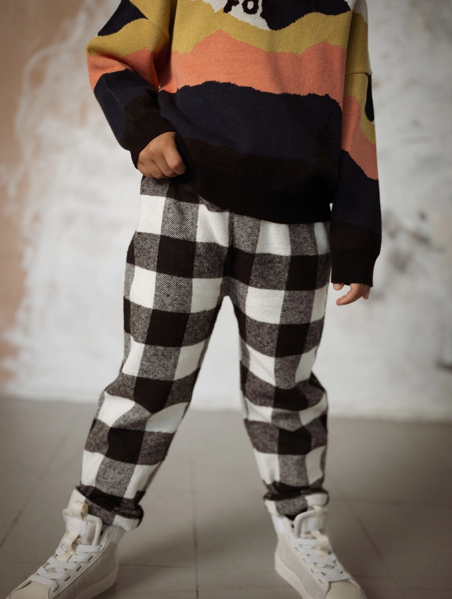 Another Fox Monochrome Flannel Check Trousers seen on a child standing on a wooden floor with a distressed background behind.
