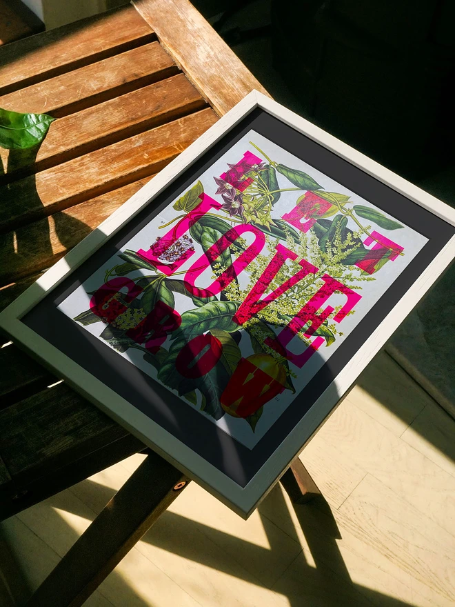 A framed limited edition artwork rests on a wooden surface, partially lit by sunlight. The letterpress print features a mix of bright pink and green leaves with the phrase "LET LOVE GROW" in bold, colorful letters rendered in fluorescent ink.