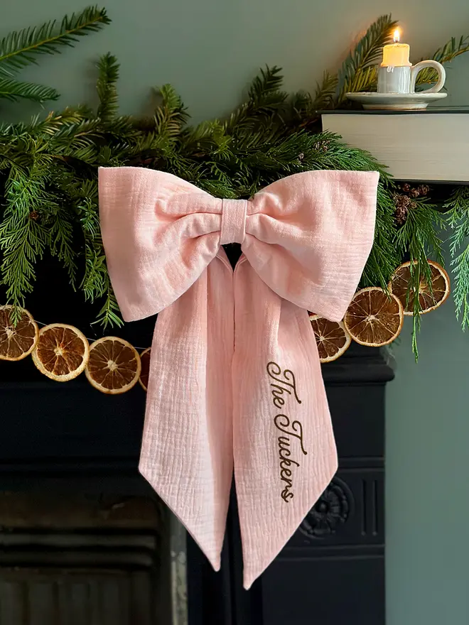A handmade Christmas bow made from blush pink cotton with the words "The Tuckers" on one tail is hung from a black mantlepiece that is decorated for Christmas with greenery, orange slice garland, and candles.