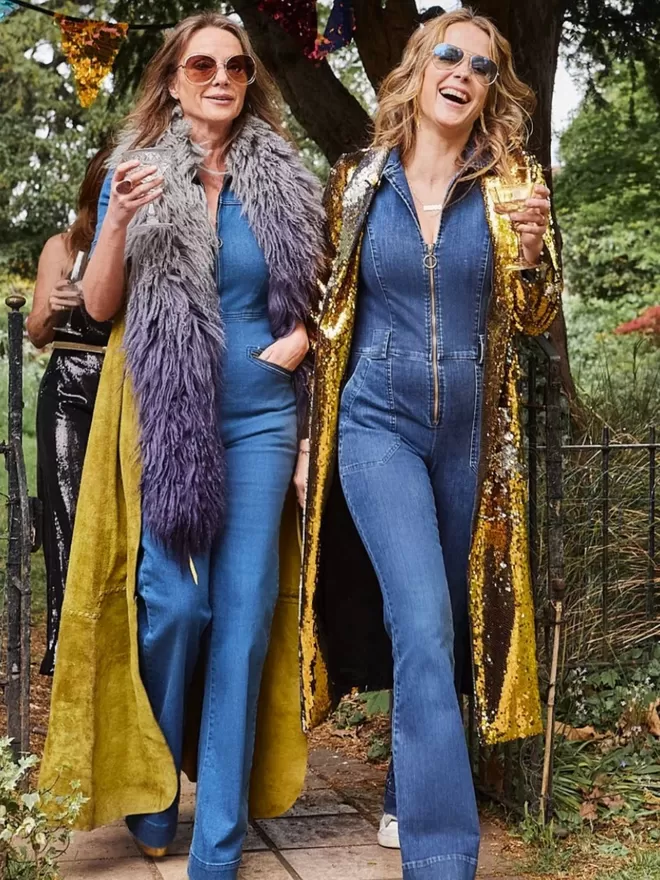 two women both wearing an anita denim jumpsuit outside in a garden wearing long coats in gold and sparkling gold 