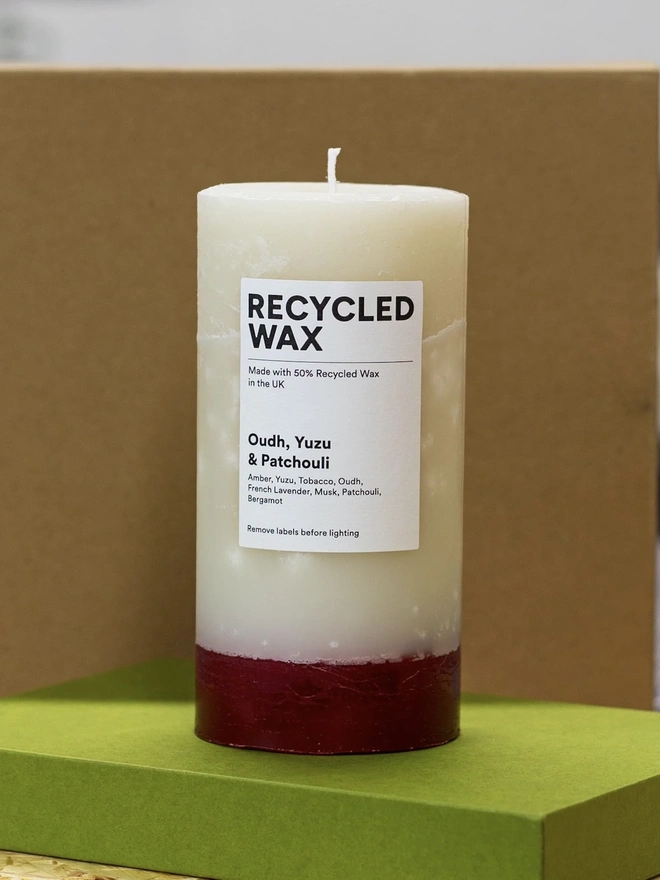 Oudh, Yuzu & Patchouli Candle (Set Of 1 Or 3 Wicks). the oudh, yuzu and patchouli candle stands on a green box against a brown background, showing its single wick and cream wax with a deep red base. a white label reads "recycled wax, made with 50% recycled wax in the uk", followed by the scent name and note list of amber, yuzu, tobacco, oudh, french lavender, musk, patchouli and bergamot. the candle's two-tone colouring and visible drip texture give it a rustic, hand-poured look.