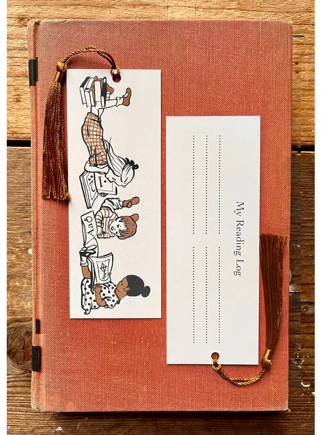a photograph of a traditional card bookmark illustrated with a vintage children's book style drawing of kids reading books. The bookmark is cream, ochre and sepia in colour, the tassel is a golden brown. Laid on a kraft envelope and there is also a gift giving label, or bookplate sticker making it a lovely alternative to a greeting card
