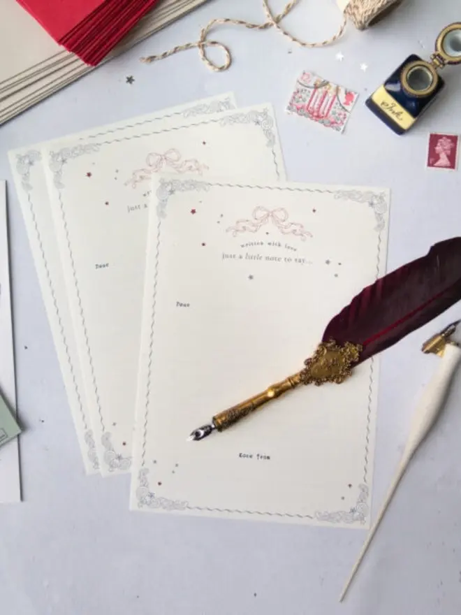 Cream lined Christmas notelets with silver scrollwork corners, a wavy border, a pink ribbon bow and scattered stars, printed "written with love, just a little note to say…" with "Dear" and "Love from". Around them are a burgundy feather quill pen, a white calligraphy pen, an ink bottle, vintage stamps, twine and red envelopes.