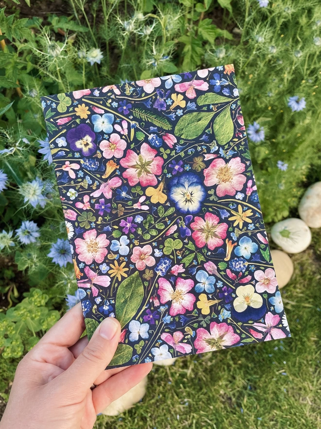 Hand Holding Botanical Print Notebook with Pretty Pressed Flower Cover, Blue Nigella Flowers in Background