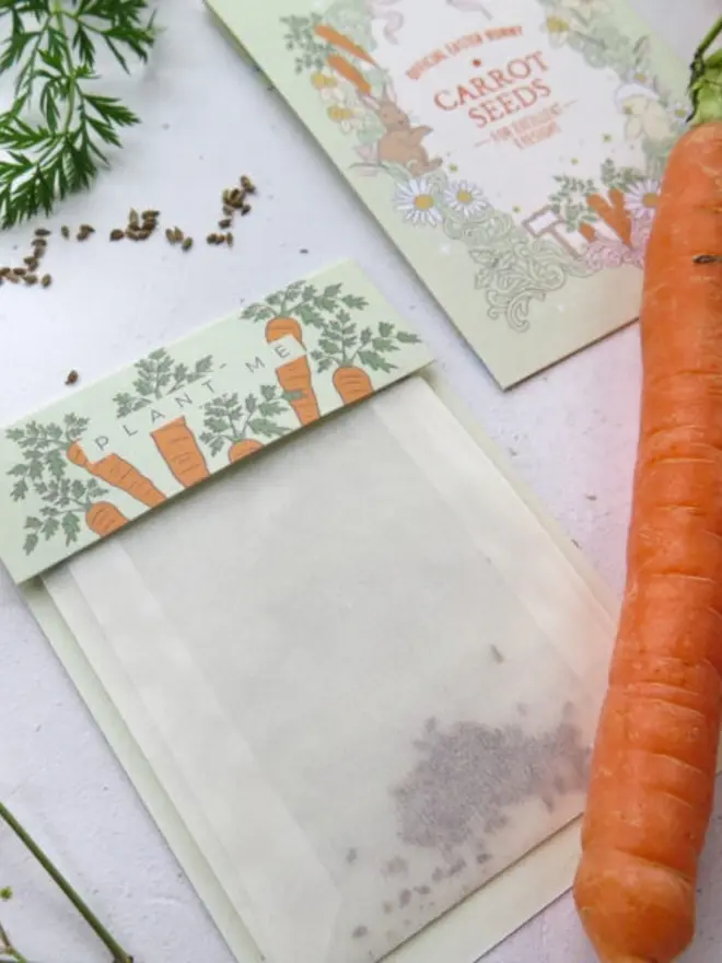Close-up of the glassine carrot seed pouch with seeds settled at the bottom. Its green topper shows illustrated carrots and the words "Plant Me."