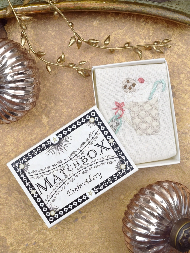 The Mug of Cocoa embroidery, inside a Matchbox displayed on a gold background with glass baubles.