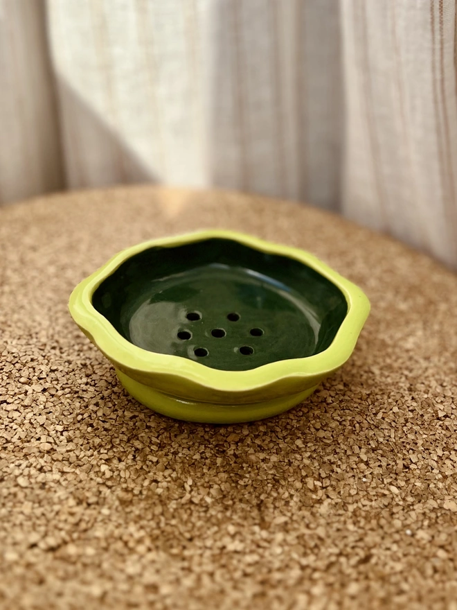 soap dish on a cork table with a fabric background. the soap dish is oval in shape with a wavy rim and has a lime green and dark green glaze