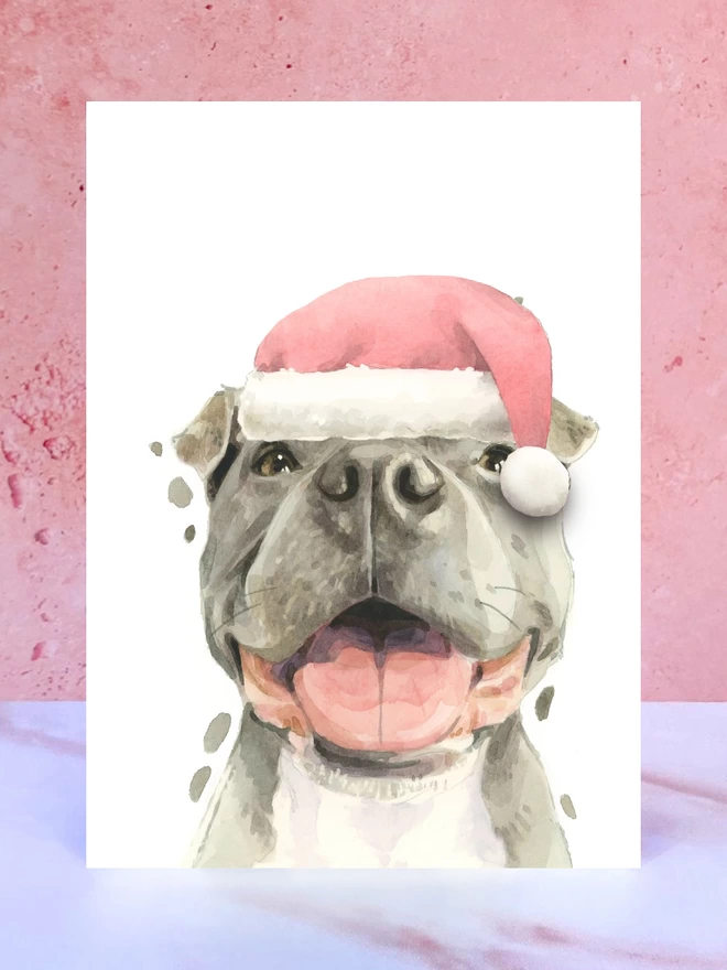 A Christmas card featuring a hand painted design of a staffordshire bull terrier, stood upright on a marble surface. 