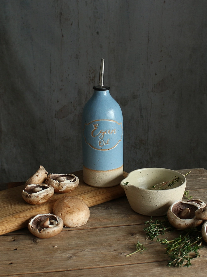 A rustic kitchen scene with a blue ceramic personalised oil pourer on a wooden table, surrounded by mushrooms, our handmade stoneware herb bowl bowl, and herbs against a dark gray wall.