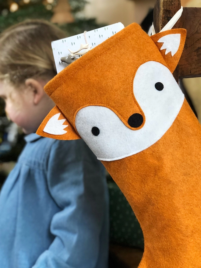A handmade felt fox stocking hangs on a wooden chair, where a little girl sits, in front of a Christmas Tree.