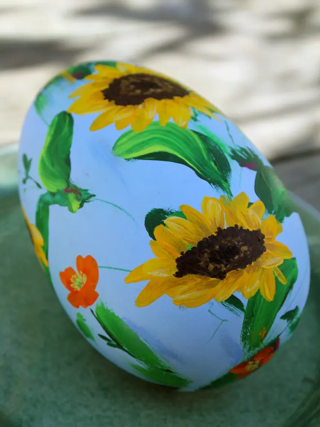 pale blue floral hand painted wooden egg, adorned in sunflowers and orange Icelandic poppy flowers, positioned on a green plate