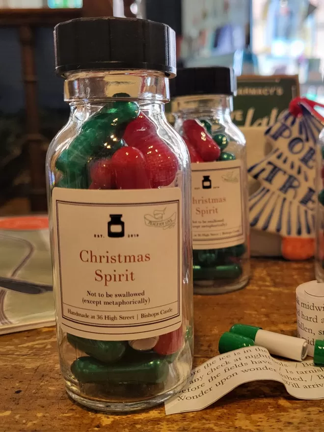 Glass bottle containing red and green Christmas Spirit poetry pills on a wooden table. Banana paper poetic extracts and pill capsules scattered about