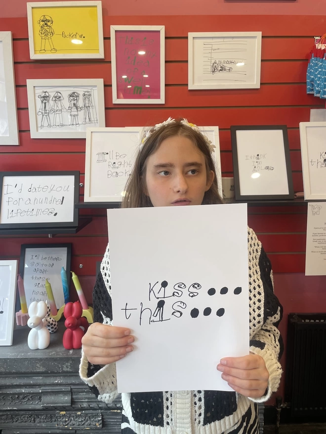 Piper a deaf, autistic teenage artist is holding her artwork up in her mums shop.  Pipers black unique handwritten font on a white background
