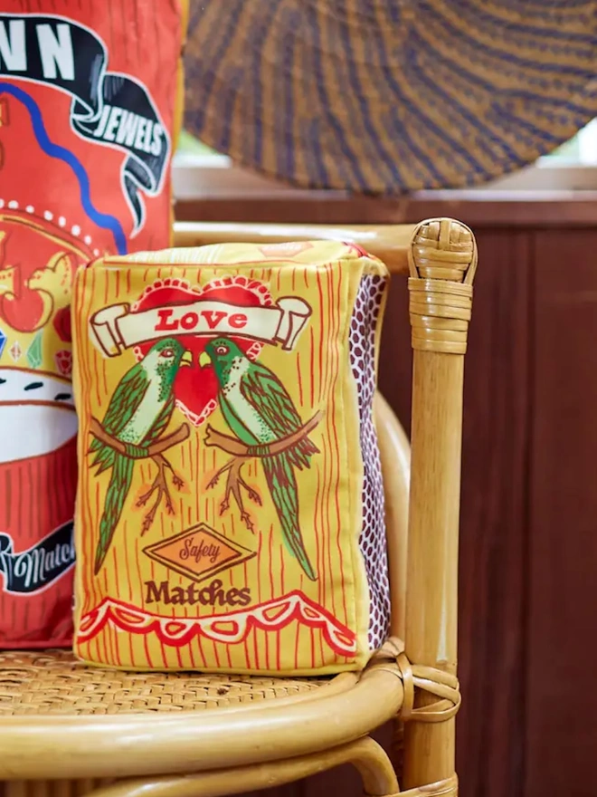 Cushion, a colourful cushion with a bird decoration sitting on a chair accompanied by another cushion. 