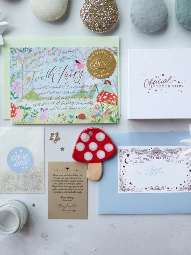 The garden notecard and white gift box sit beside a blue envelope addressed to Alfie. A red ceramic toadstool, a Magic Dust packet and the Tooth Fairy's note are laid out below.