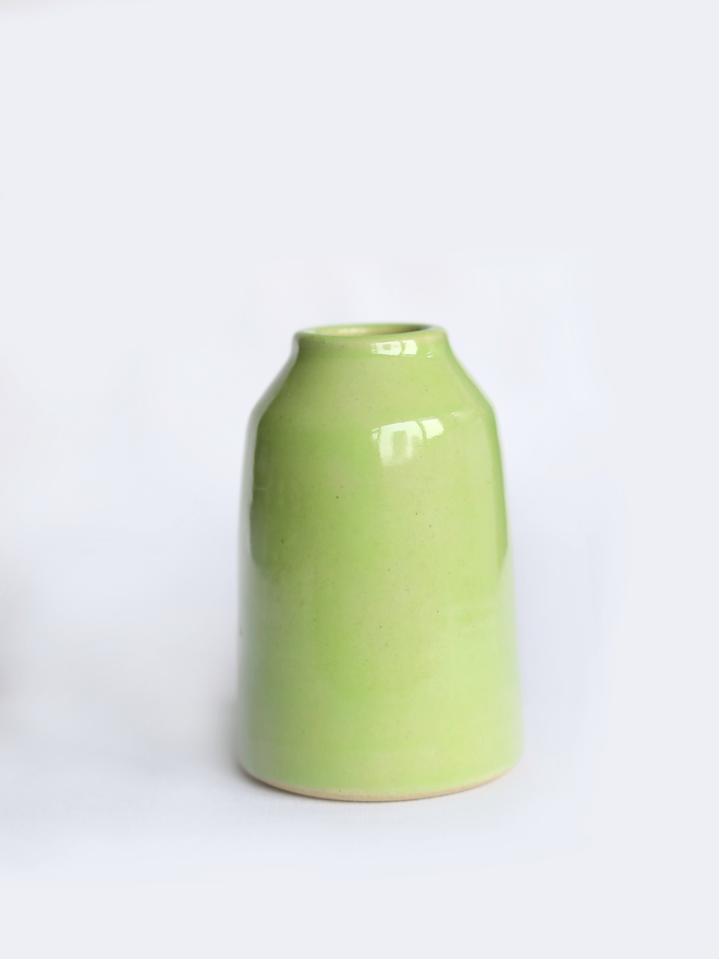 A small handmade ceramic bud vase in glossy spring green glaze, with a rounded bottle shape and narrow neck, photographed against a plain white background