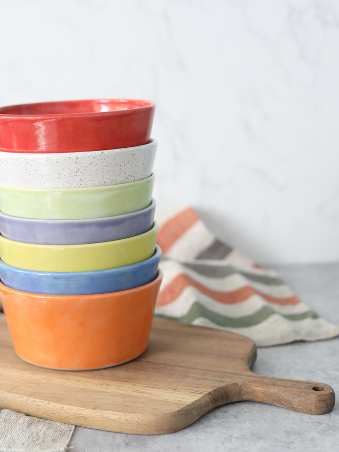 A tall stack of seven ceramic breakfast bowls in rainbow colours — red, speckled white, green, blue, orange, yellow, and purple — stacked on a wooden chopping board, with a striped linen cloth beside them.