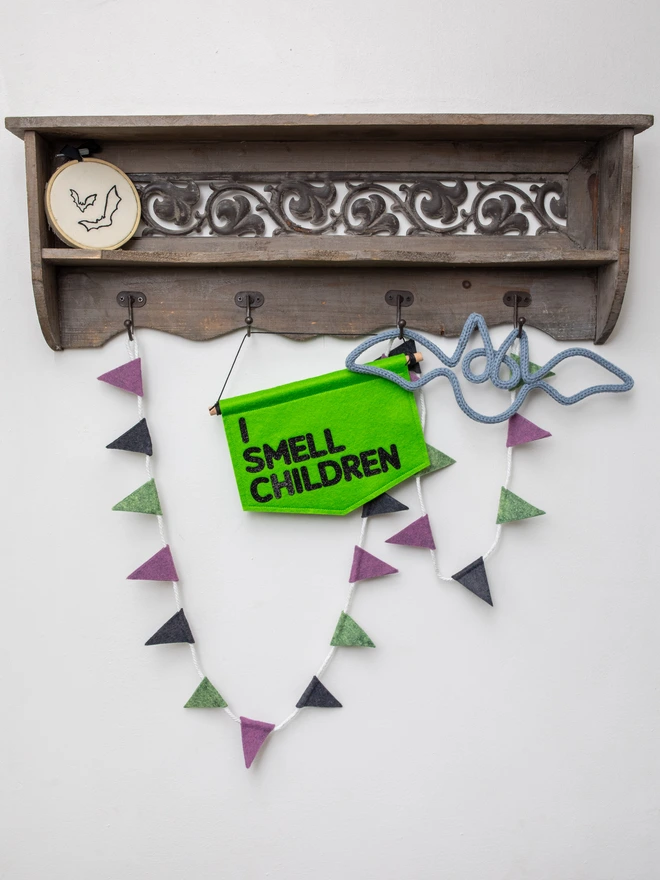 Green, fell halloween banner that has "I SMEEL CHILDREN" written on it in black glitter felt. The banner is hanging from a wooden shelf unit and surrounded by halloween decor