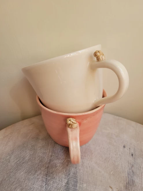 a pink ceramic cup is on a wooden tray with a white cup stacked on top both have a miniature beige lop bunny rabbit attached to the top of the handle and tiny pawprints on the cup