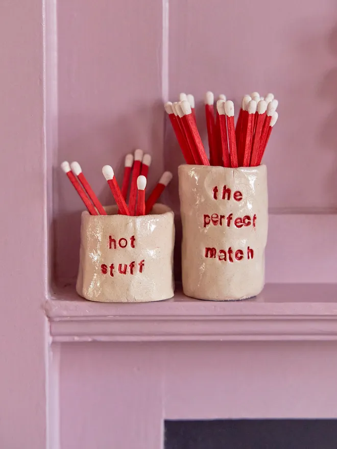 Two ceramic match holders, one short, one tall on a pink shelf