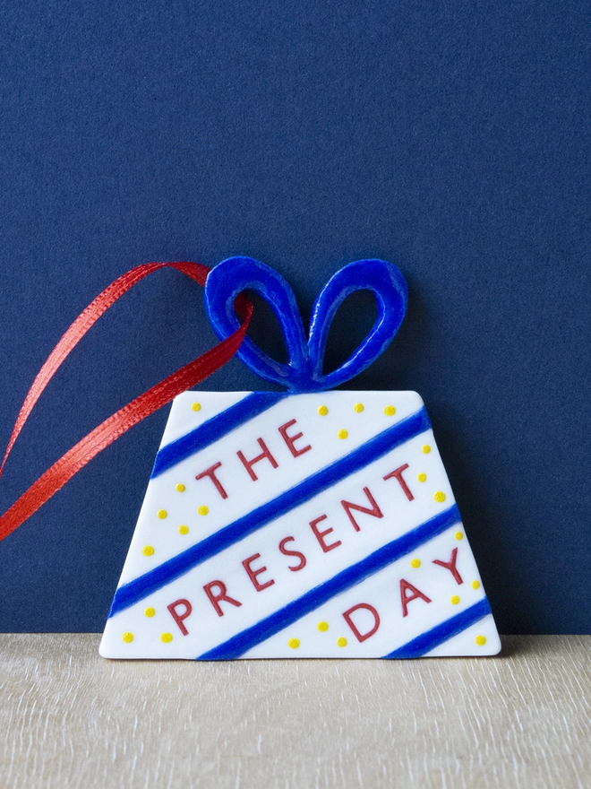 Ceramic porcelain Christmas decoration with The Present Day written on the side. A white present-shaped Christmas decoration with red lettering, decorated with blue stripes, yellow spots and a red satin ribbon.